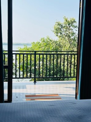 A room with a view of a body of water and trees, a balcony with a railing, and a patterned rug on the floor. - cormorant bath (Rekawa)
