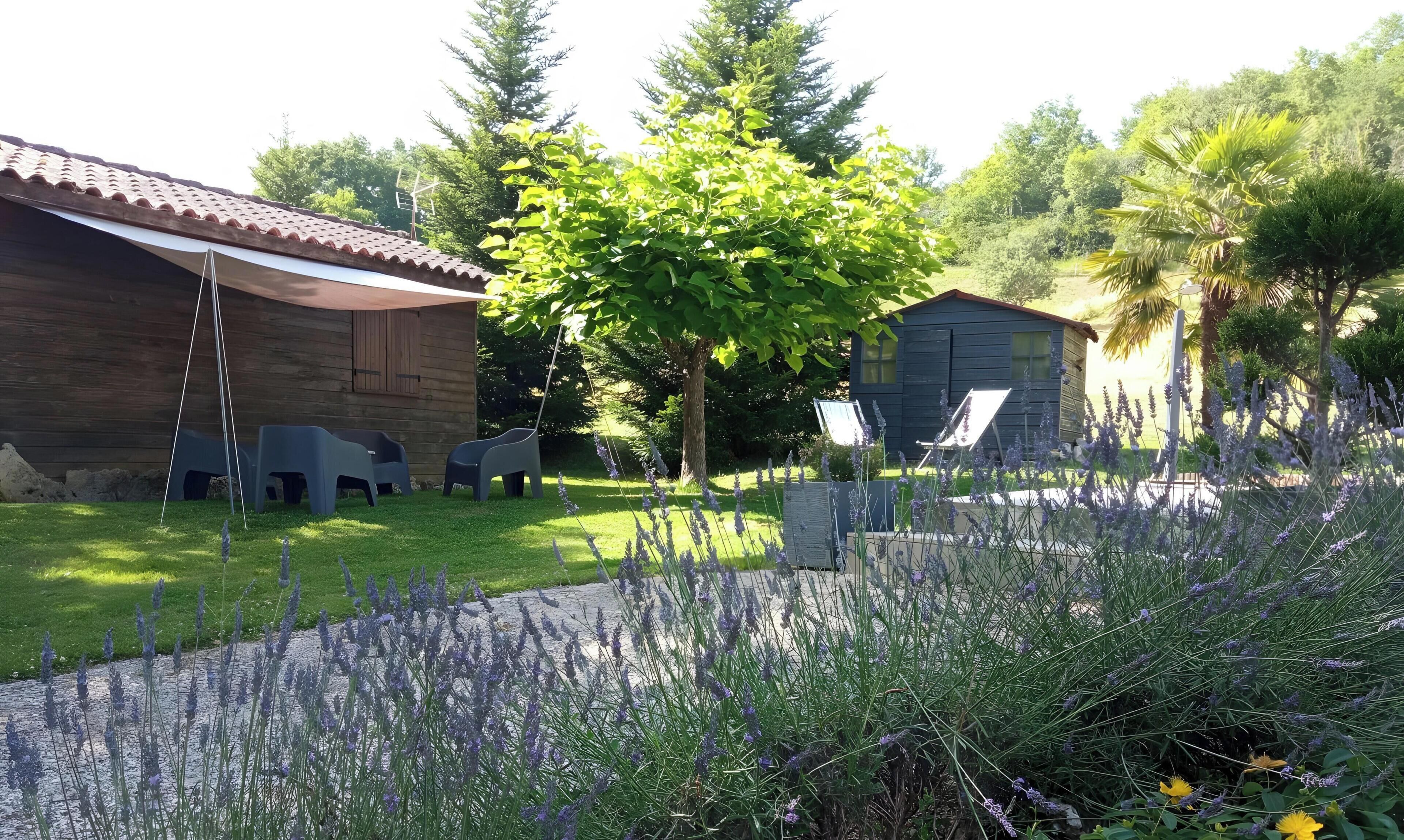 Un bâtiment en bois avec un toit en tuiles, deux chaises en plastique, un arbre et un jardin agrémenté de lavande.