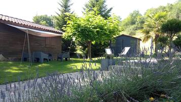 Un bâtiment en bois avec un toit en tuiles, deux chaises en plastique, un arbre et un jardin agrémenté de lavande.
