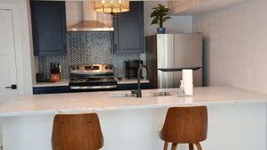A modern kitchen with dark cabinets, a stainless steel refrigerator, a built-in oven, and a bar area with two wooden stools. - Waterfront Coastal Escape (Atlantic Beach)
