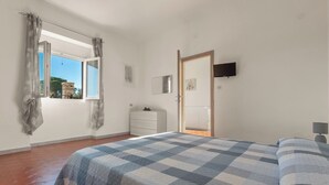 Quarto com colcha xadrez, penteadeira branca, televisão na parede e janela com cortinas.