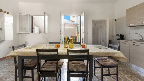 Uma sala de jantar com uma mesa posta para quatro pessoas, cadeiras com almofadas, uma cozinha com armários e uma porta aberta para outro cômodo.