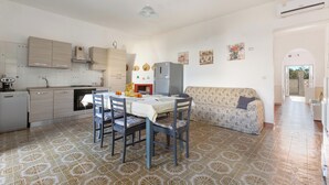 Uma cozinha com mesa de jantar, cadeiras e um sofá. Há uma geladeira, micro-ondas e pia. O chão tem azulejos estampados.