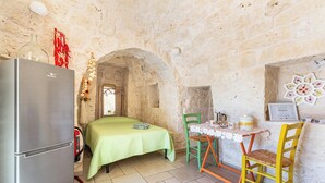 A stone-walled room with a bed, a table with chairs, a refrigerator, and a small table with a vase. - Beltrullo Pietrevive by BarbarHouse (Martina Franca)