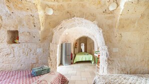 A rustic room with a stone wall, a bed with a patterned bedspread, a green table with a cloth, and a refrigerator. - Beltrullo Pietrevive by BarbarHouse (Martina Franca)