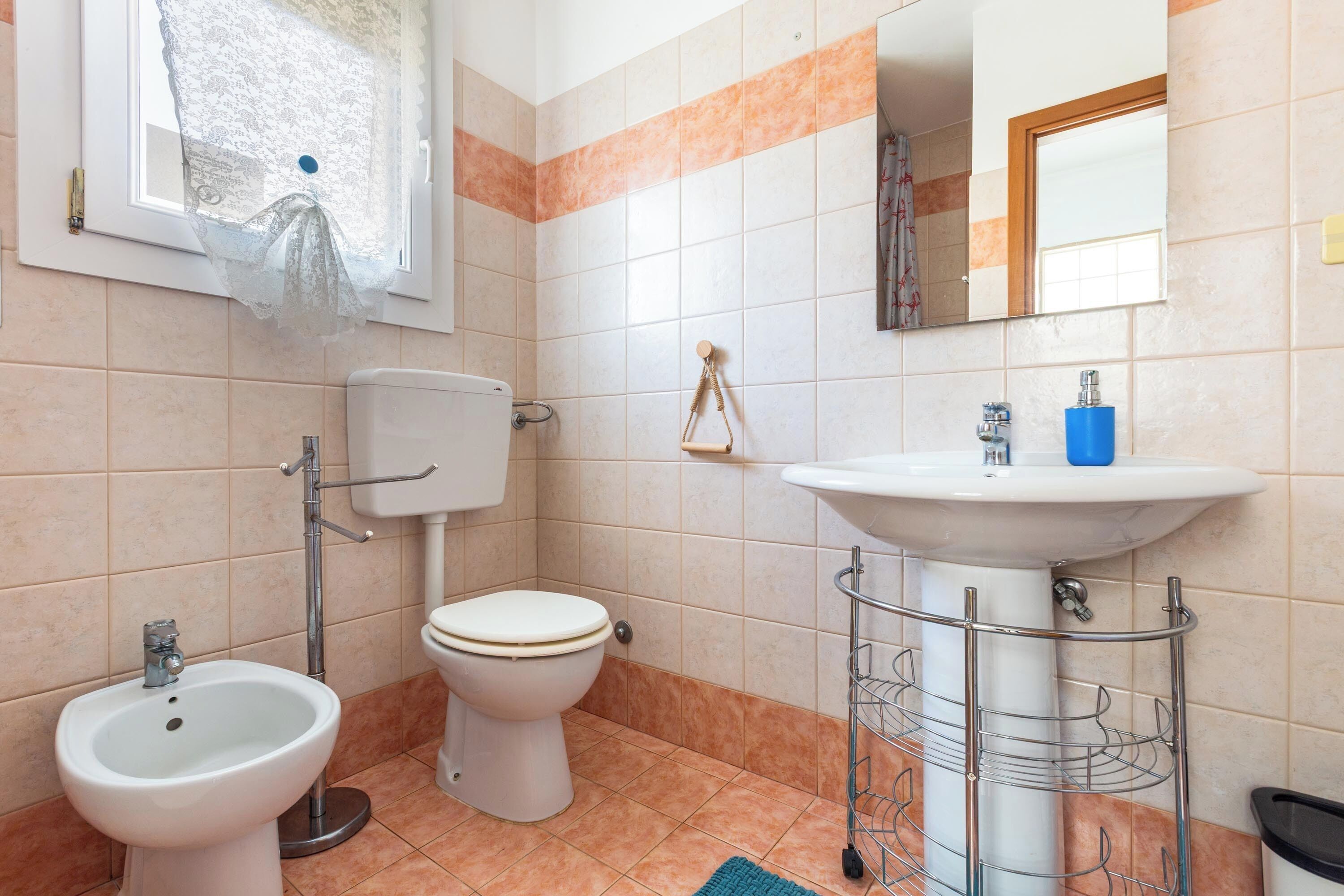 Un baño con inodoro, bidé, lavabo y ventana con cortina de encaje.