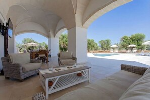 Un espacio de estar al aire libre techado con mobiliario de mimbre, piscina y sombrillas.