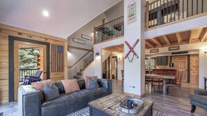 Ein großzügiger Wohnbereich mit einem schwarzen Ledersofa, einem Couchtisch und einem gemusterten Teppich. Eine Treppe führt zu einer zweiten Etage mit einem Balkon.