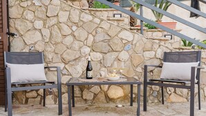 Un muro de piedra, dos sillas de metal con cojines, una mesa con una botella, un cuenco y un vaso.