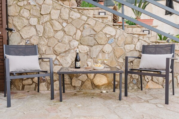 A stone wall, two metal chairs with cushions, a table with a bottle, a bowl, and a glass.