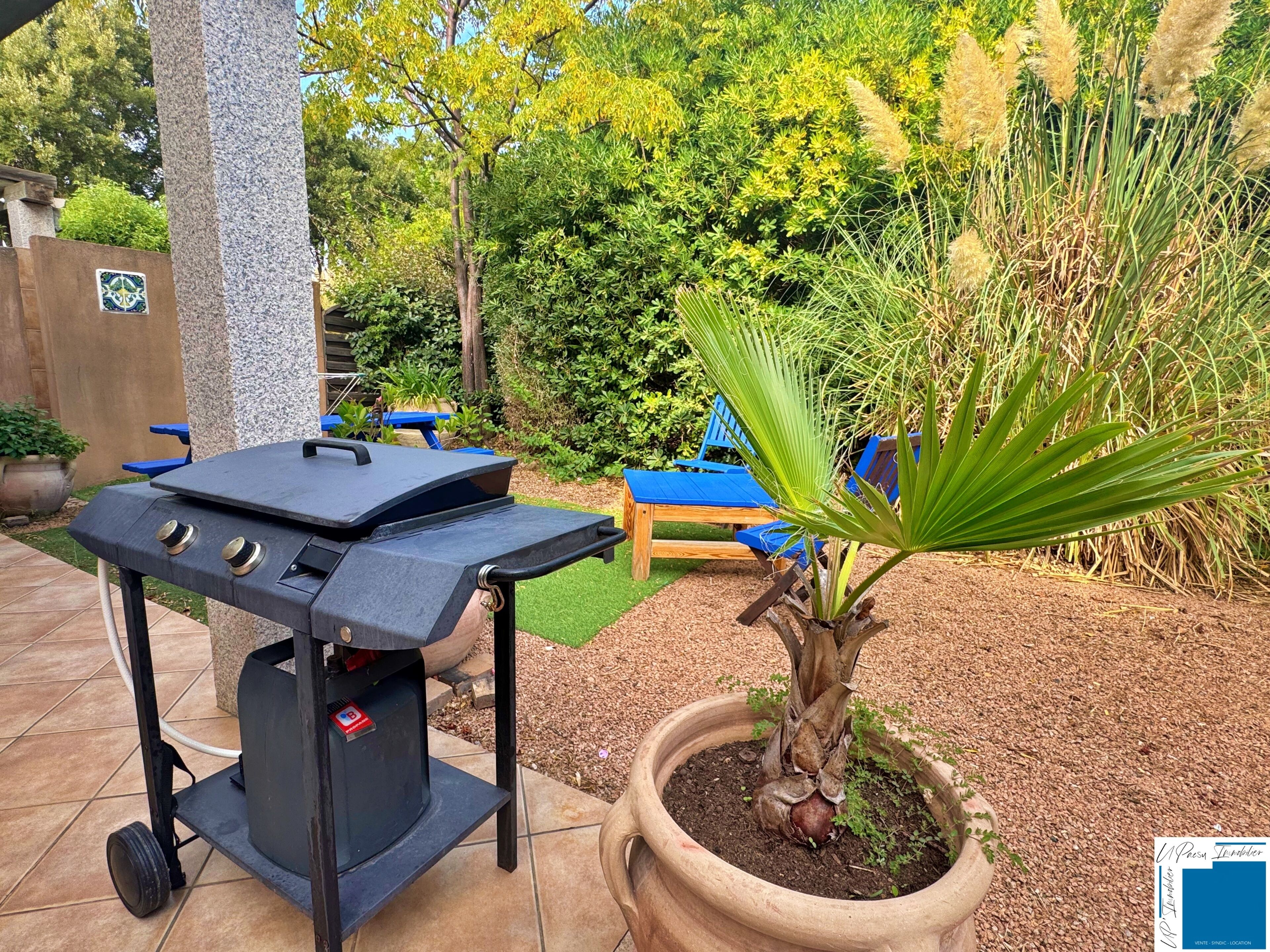A barbecue grill with a control panel, a potted plant, and a blue lounge chair in a garden setting.