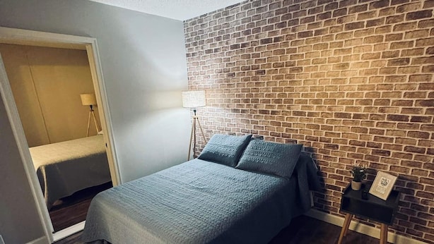 A bedroom with a brick-patterned wall, a bed with a grey bedspread, a small bedside table with a lamp, and a wooden floor.