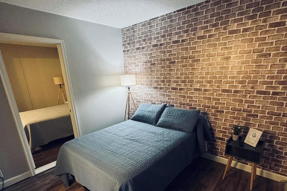 A bedroom with a brick-patterned wall, a bed with a grey bedspread, a small bedside table with a lamp, and a wooden floor.