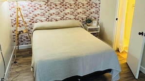 A bedroom with a brick-patterned wall, a bed with a grey blanket, a bedside table with a plant, and a lamp.