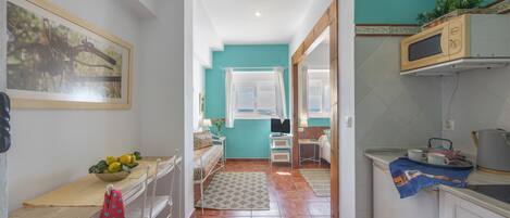 Un estrecho pasillo conduce a un dormitorio con cama, un escritorio con un frutero y una ventana con cortinas.
