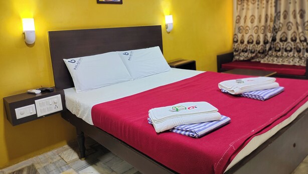 Habitación de hotel con cama, almohadas, mesita de noche, lámpara de pared, un cuadro y ventana con cortinas.