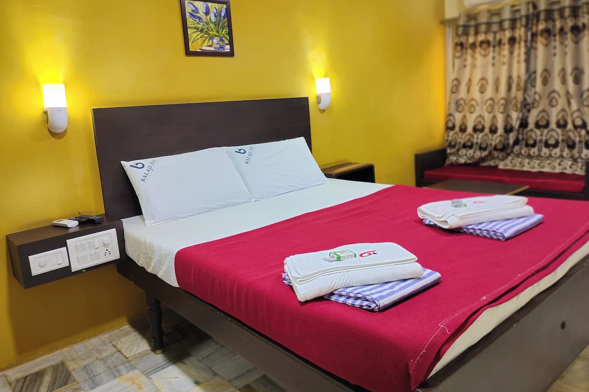 A hotel room with a bed, pillows, a nightstand, a wall-mounted light, a painting, and a window with curtains.