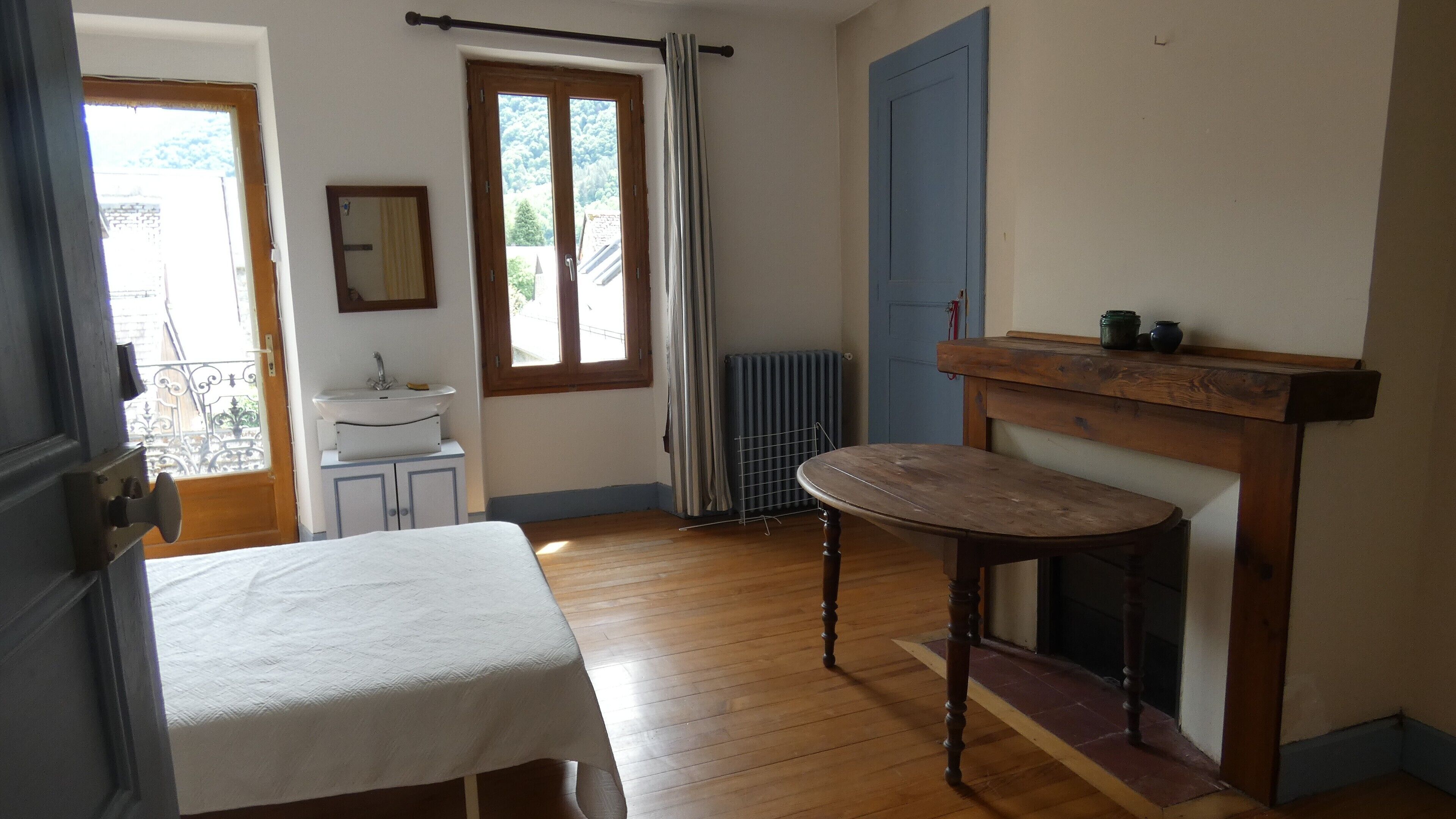 Ein Zimmer mit einem Bett, einem Tisch, einem Heizkörper, einem fenster mit Vorhängen, einer Tür und einem kleinen Schrank mit Spiegel.
