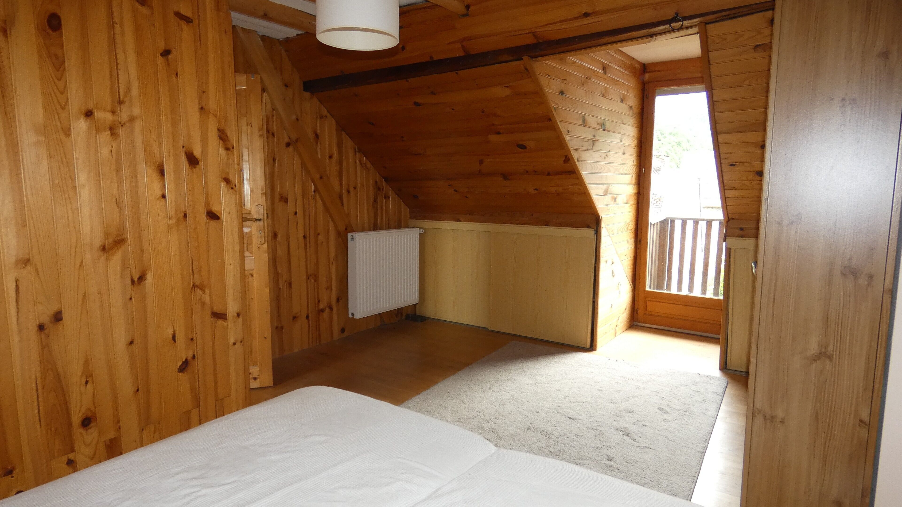 Ein Zimmer mit Holzwänden und Schrägen, einem Heizkörper und einem Blick durch eine offene Tür auf einen Außenbalkon.