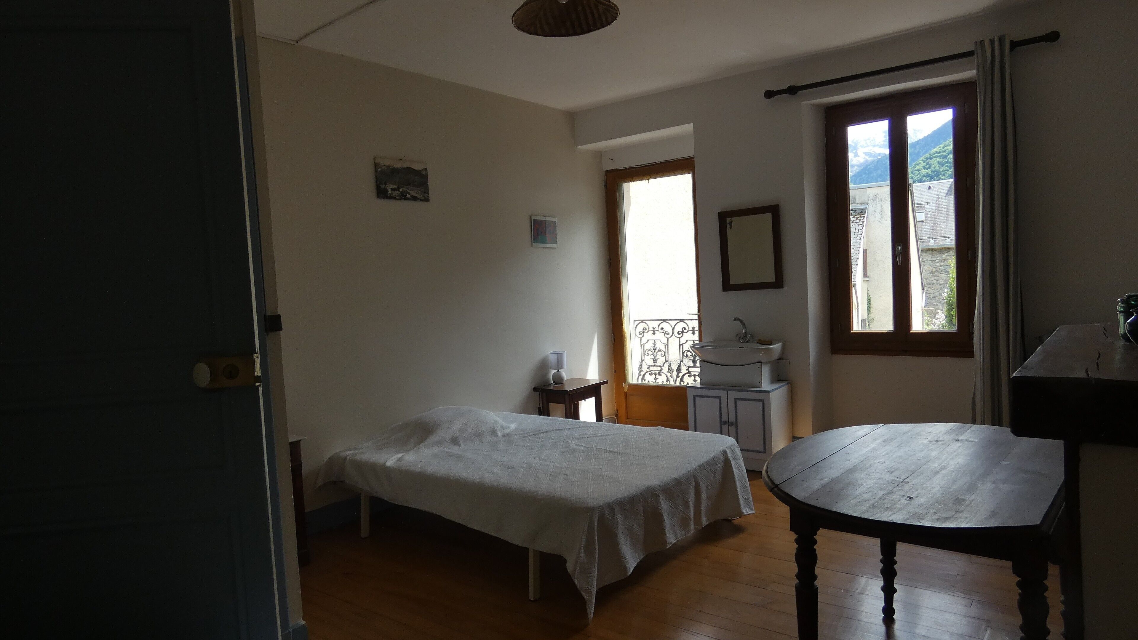 Quarto com cama, mesa de cabeceira pequena, janela com vista, banheiro com pia e uma cadeira de madeira.