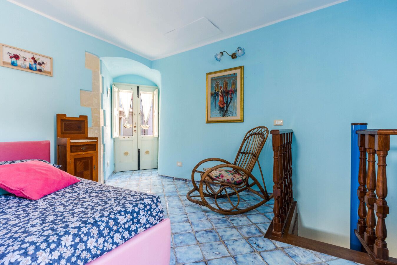 A bedroom with a bed, a rocking chair, a door, and a wall-mounted picture.