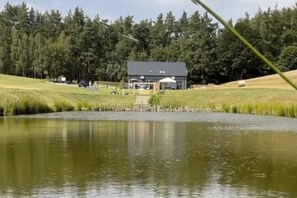 A building with a large open garage area, surrounded by a grassy field and a body of water. - Large Holiday Home With a Jacuzzi on the Terrace (Koscierzyna)
