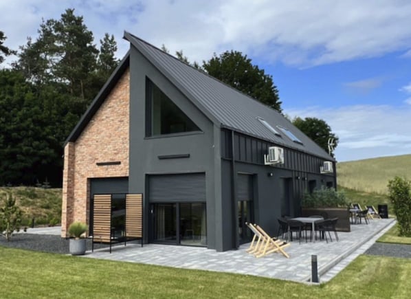 Ett modernt hus med ett sluttande tak, stora fönster och en uteplats med utemöbler.