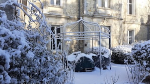A snow-covered garden with a metal gazebo, stone building with multiple windows, and snow-laden bushes. - LE GITE DU CHATEAU NEAR MARAIS POITEVIN (Fors)