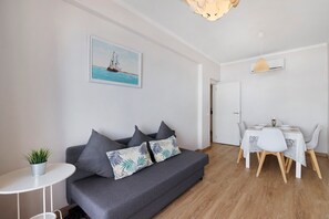 A modern living room with a grey sofa, a dining table with white chairs, a small potted plant, and a painting of a sailboat on the wall. - Apartment 'Jc Mar Ll - 2 Min To The Beach' with Balcony, Wi-Fi and Air Conditioning (Albufeira)