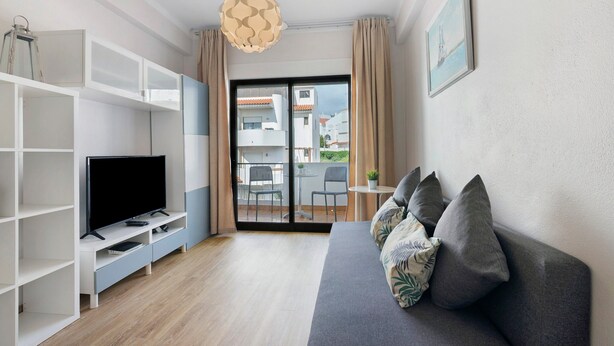 Un soggiorno con divano, TV e scaffali, un balcone con sedie e vista sugli edifici.