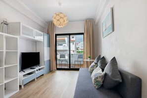Ein Wohnzimmer mit Sofa, Fernseher und Regalen, ein Balkon mit Stühlen und Blick auf Gebäude.