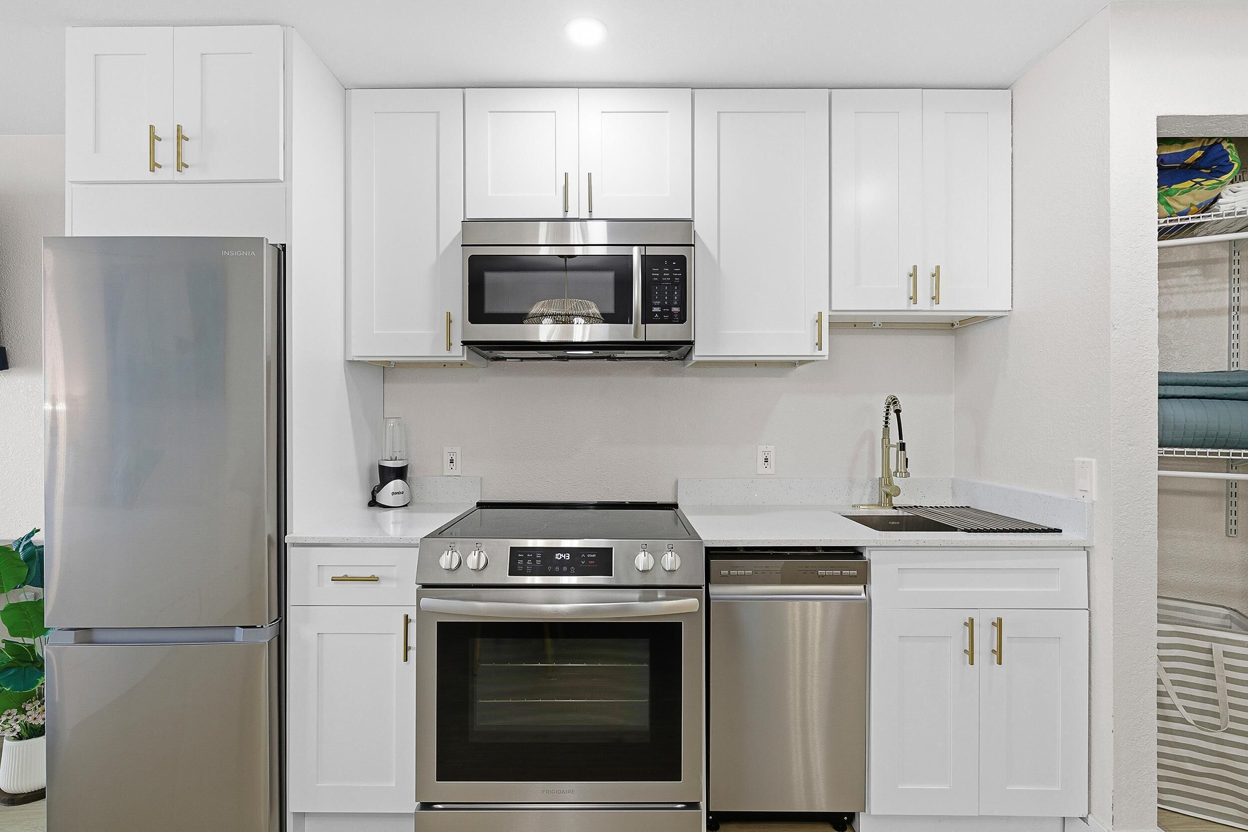 A compact kitchen with a refrigerator, microwave, oven, and dishwasher, all in stainless steel, with white cabinetry and gold handles.