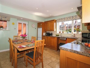 A kitchen with wooden cabinets, a dining table with chairs, a clock, and a window with a view of a house. - The Rumbles (Ludlow)