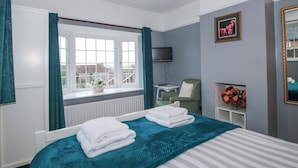 Un dormitorio con cama, ventana con cortinas, una silla, televisor y un cuadro en la pared.