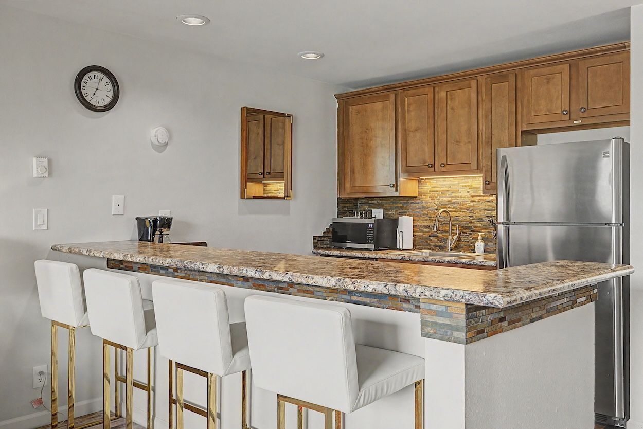 A kitchen with a granite countertop, wooden cabinets, a stainless steel refrigerator, and a clock on the wall.