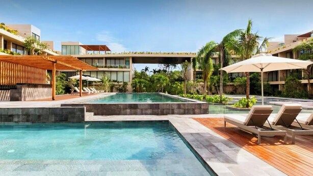 Piscina con sillones de descanso y sombrillas, rodeada de follaje y edificios modernos.