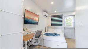 A bedroom with a bed, a desk, a chair, a TV, and a window with blinds. - The Reef in Fort Lauderdale (Fort Lauderdale)