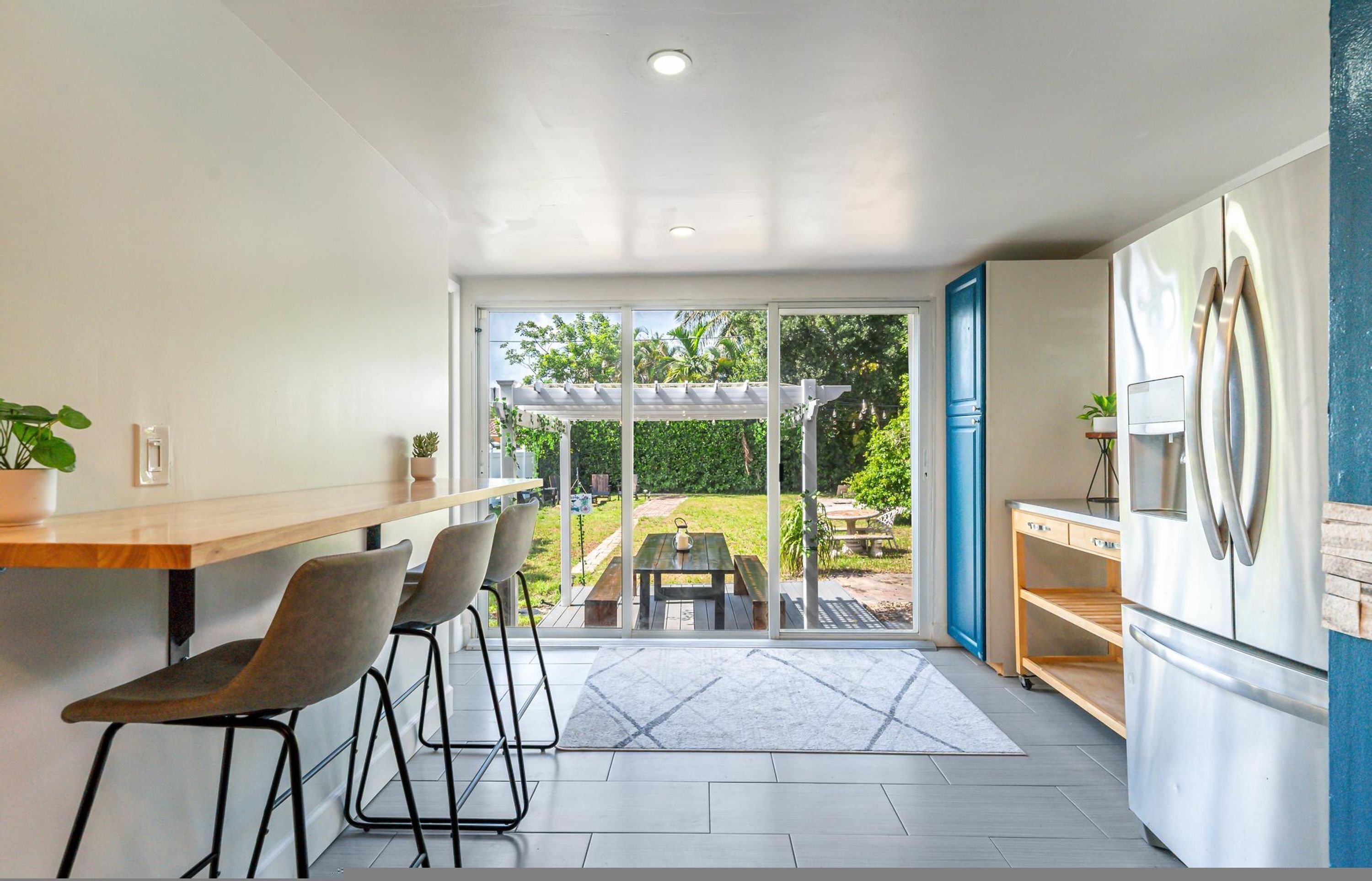 A modern kitchen with a large refrigerator, bar stools, a dining table, and a view of a backyard with a pergola.