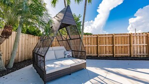 Un espace extérieur aménagé en forme de triangle, avec un banc rembourré et un dossier en osier, entouré d’une clôture en bois et de palmiers.