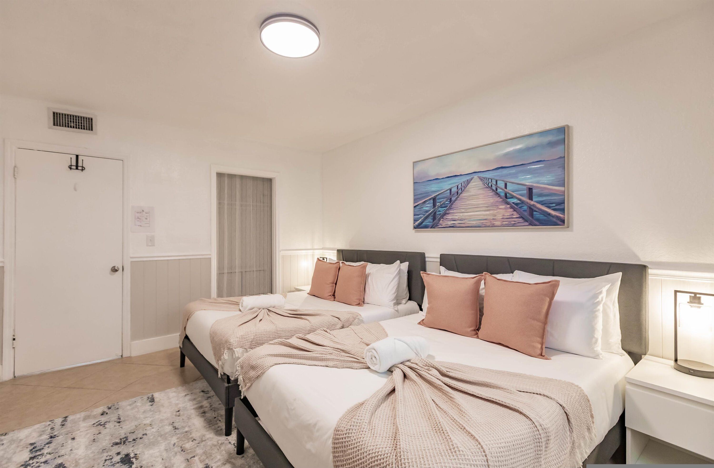 A hotel room with two beds, a painting of a pier, a white door, and a bedside lamp.
