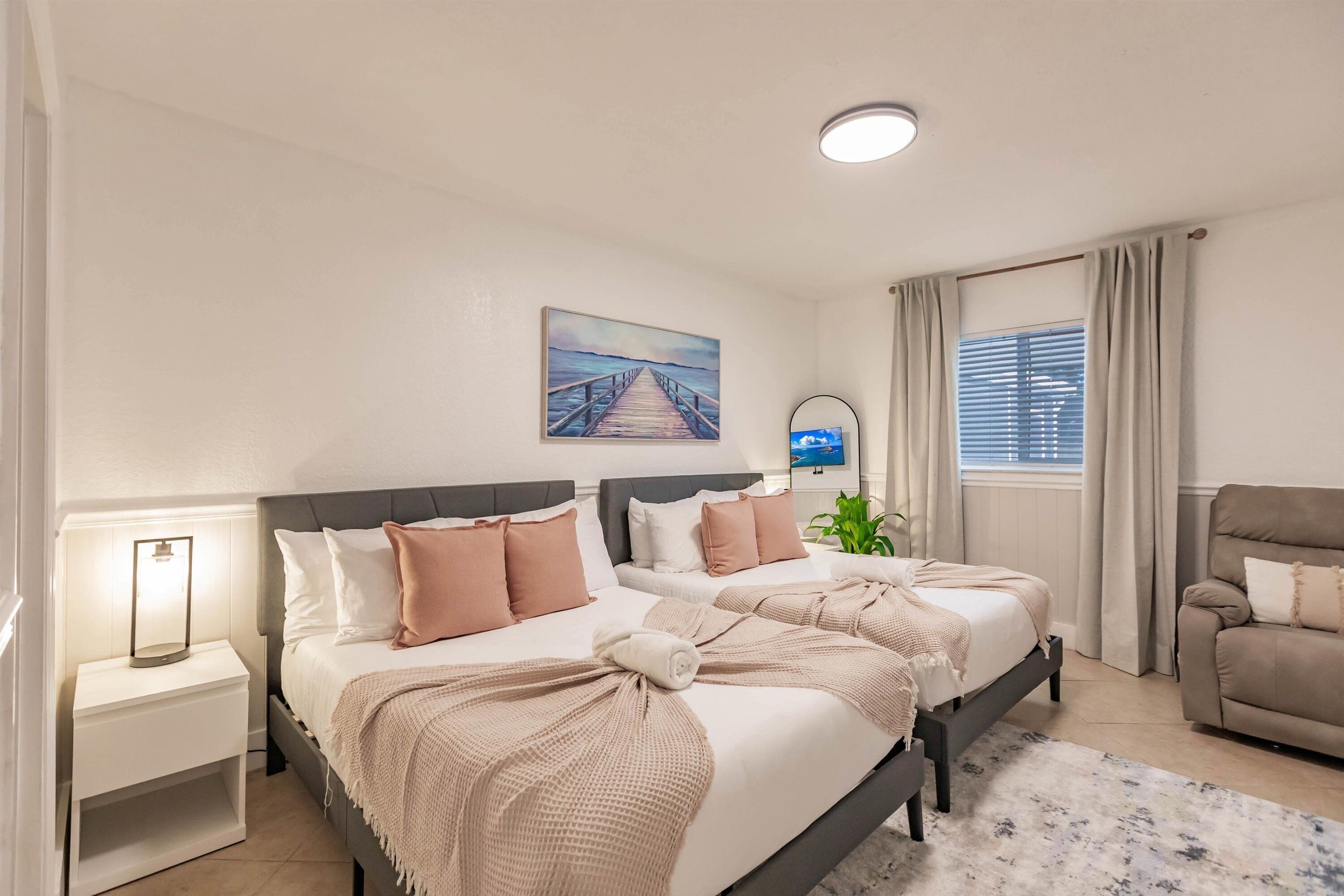 Ein Schlafzimmer mit zwei Betten, einem Nachttisch, einer Lampe, einem Gemälde, einem Fenster mit Vorhängen und einem Sofa.