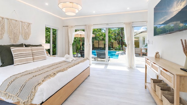 A bedroom with a large bed, a wooden bedside table, a ceiling light, and a view of an outdoor pool area. - Fox Pearl 4Bd Pool House- Las Olas Luxury Getaway (Fort Lauderdale)