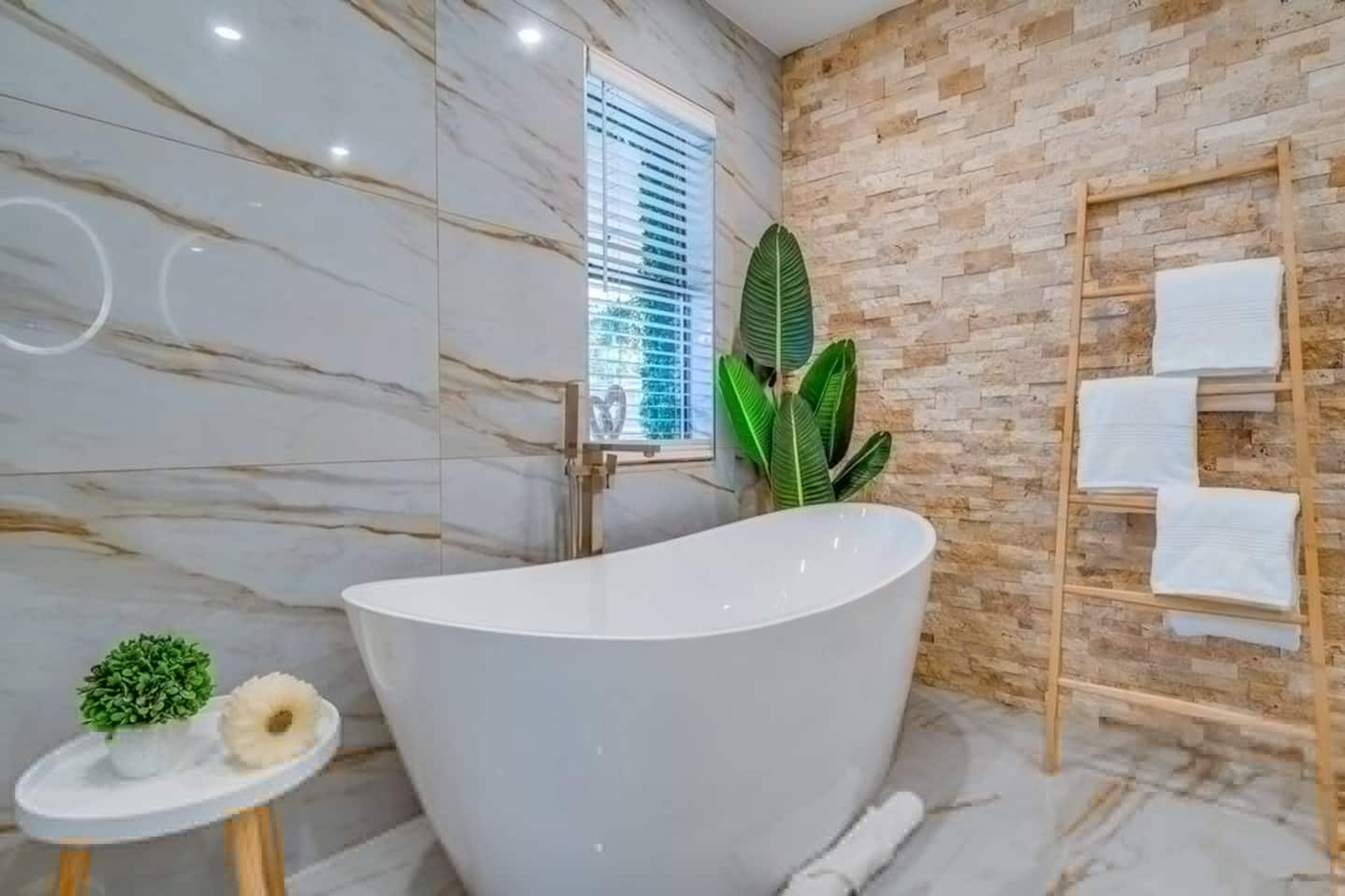Ein modernes Badezimmer mit einer freistehenden Badewanne, einer Holzleiter mit Handtüchern, einem Topf mit Pflanze und einem kleinen Tisch mit Pflanze und einem dekorativen Gegenstand.