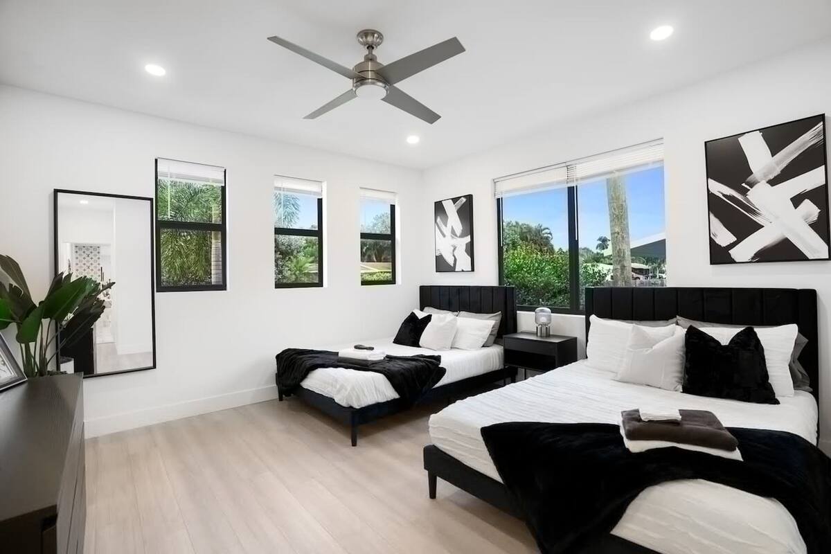 A modern bedroom with two beds, large windows, ceiling fan, and artwork.