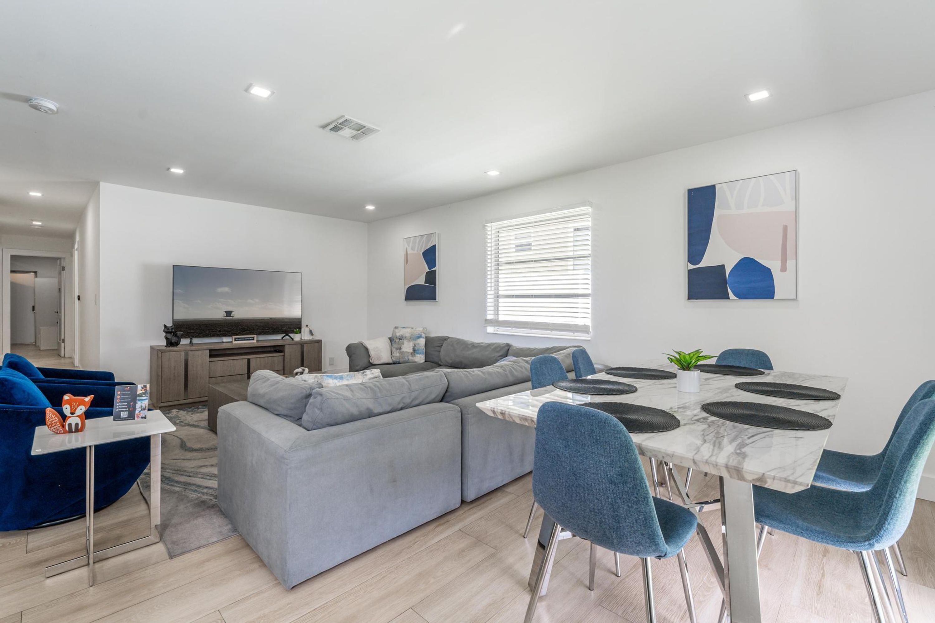 A modern living room with a sectional sofa, a dining table with chairs, a flat-screen TV, and abstract wall art.