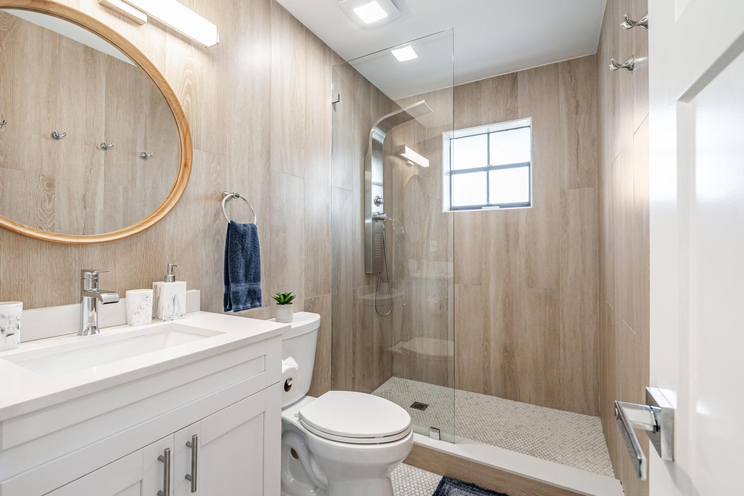 A modern bathroom with a wooden vanity, a toilet, a shower with a glass door, and a small window.