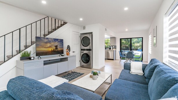A modern living room with a sofa, a coffee table, and a TV. There is a staircase with a railing, a washing machine, and a kitchen area visible in the background. - Fox Palace Pool House (Fort Lauderdale)