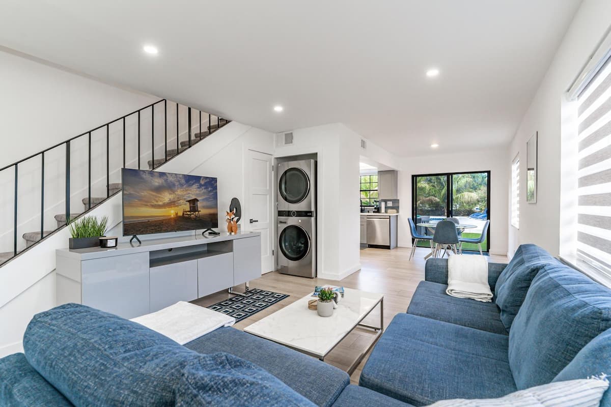 A modern living room with a sofa, a coffee table, and a TV. There is a staircase with a railing, a washing machine, and a kitchen area visible in the background.