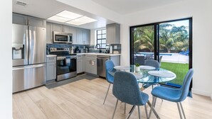 A modern kitchen with stainless steel appliances, a dining area with blue chairs, and a large sliding glass door to a garden. - Fox Palace Pool House (Fort Lauderdale)