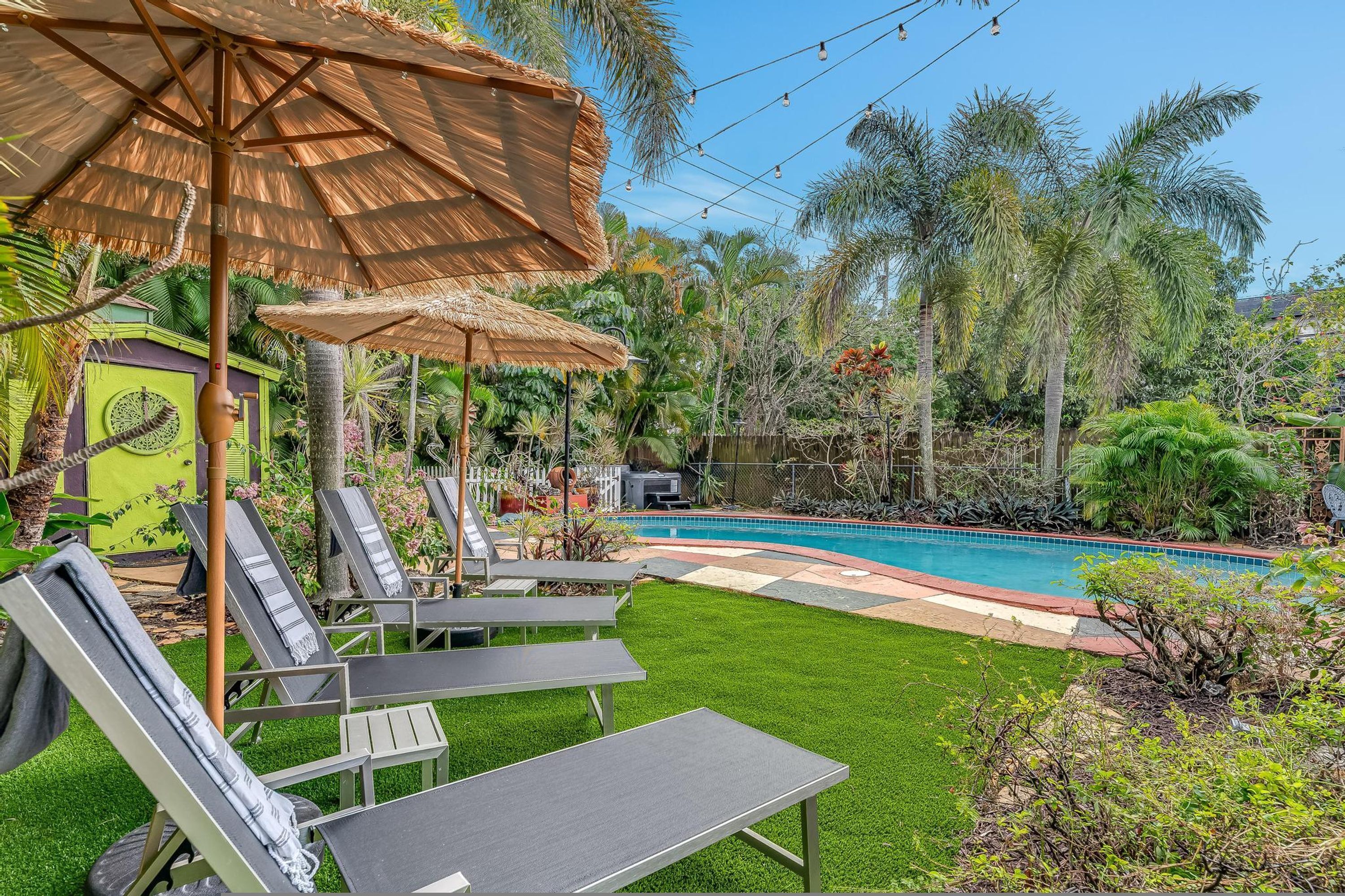 Un jardin avec une piscine, des chaises longues et un parasol en chaume.